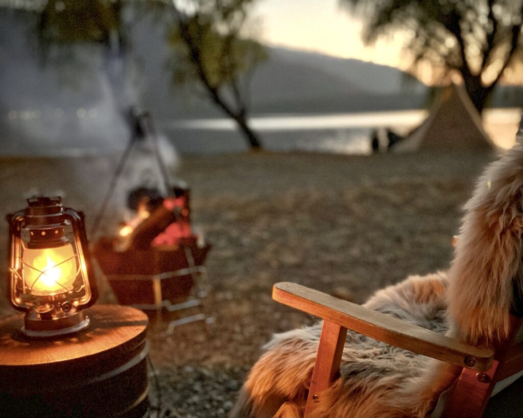 夕暮れのキャンプ場でテーブルに置かれたオイルランタンとムートンラグを敷いた木製チェア