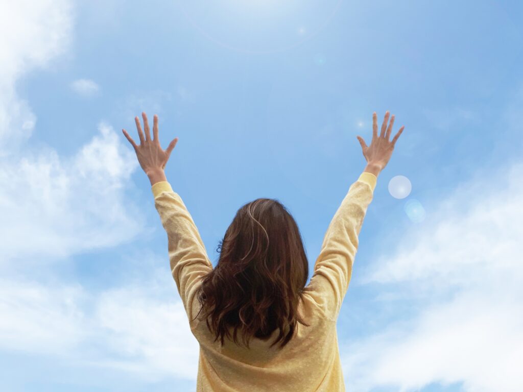 青空に向かって両手を高く広げて深呼吸する女性の後ろ姿とリフレッシュした様子