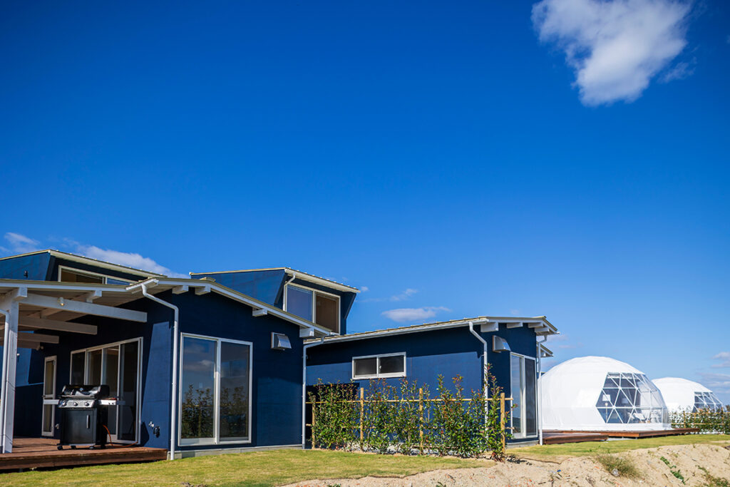 青空に映える青いヴィラ（コテージ）とドームテントが並ぶ海辺のグランピング施設