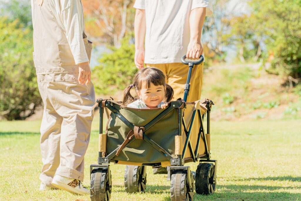 明るい芝生の上で、大人に引かれたアウトドアワゴンに乗って笑顔を見せる小さな女の子