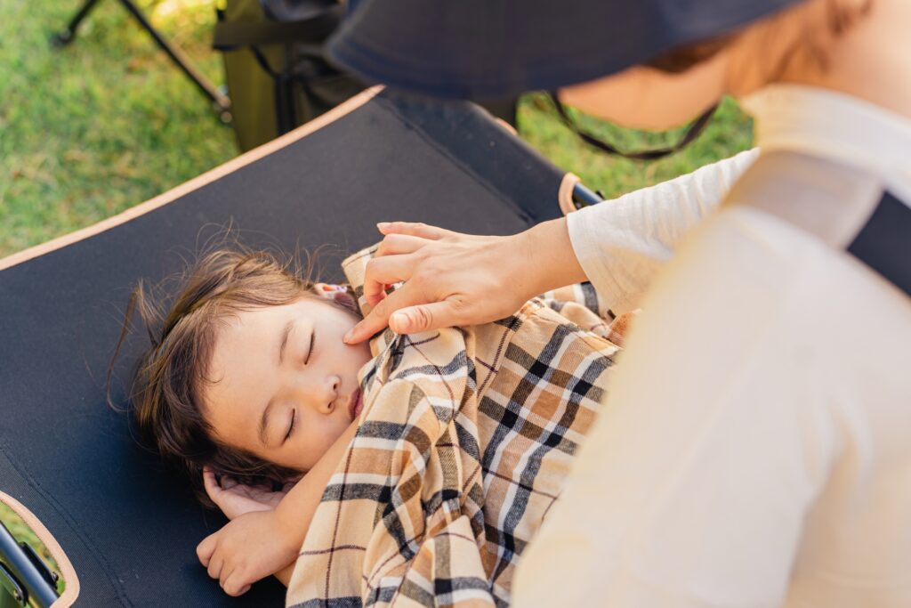 キャンプ用コットで眠る子どもに、優しくチェック柄の毛布（上着）をかける母親